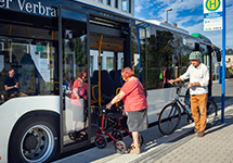 Ältere Frau mit Rollator steigt in einen Linienbus an einer Haltestelle ein, Mann mit Fahrrad wartet daneben.