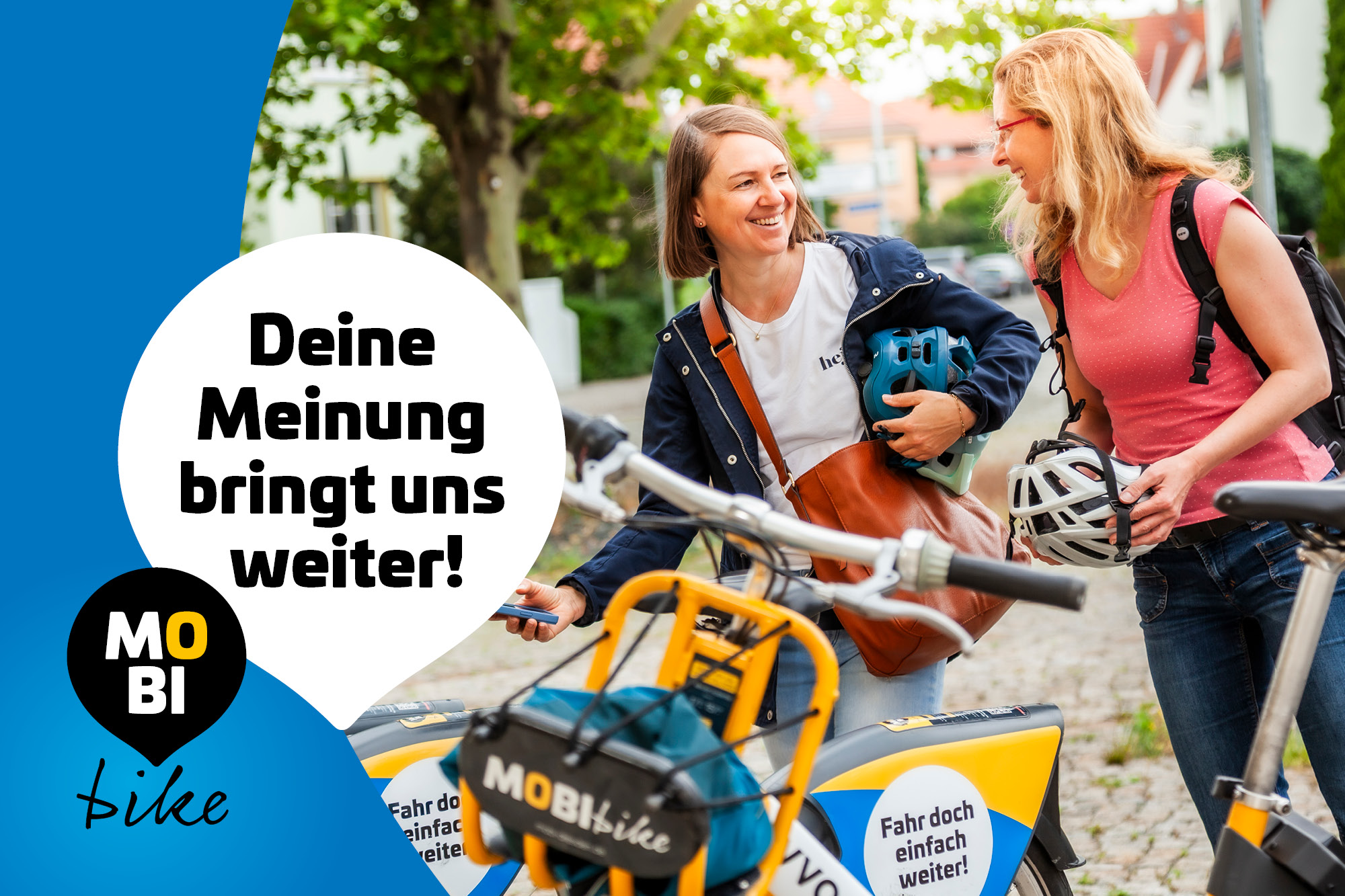Zwei junge Frauen mit Fahrradhelmen in der Hand stehen neben gelben MOBIbike-Leihrädern auf einem Gehweg, im Hintergrund Bäume und Gebäude.